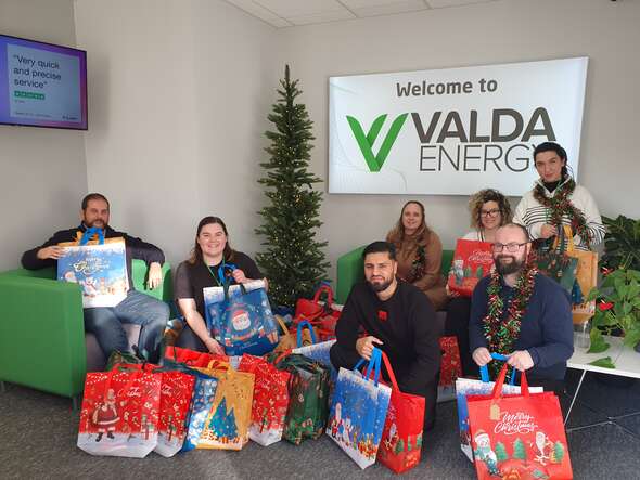 People from across Valda, holding the gift bags donated to the Bicester Baby Bank Appeal in front of a lit-up Christmas tree. 